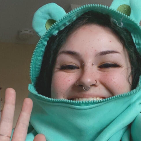 Fae is a white person with multiple facial piercings. They are smiling, wearing a frog hoodie, and have makeup on.